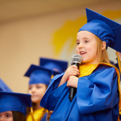 Pre-K student speaking into microphone with blue graduation cap and gown.