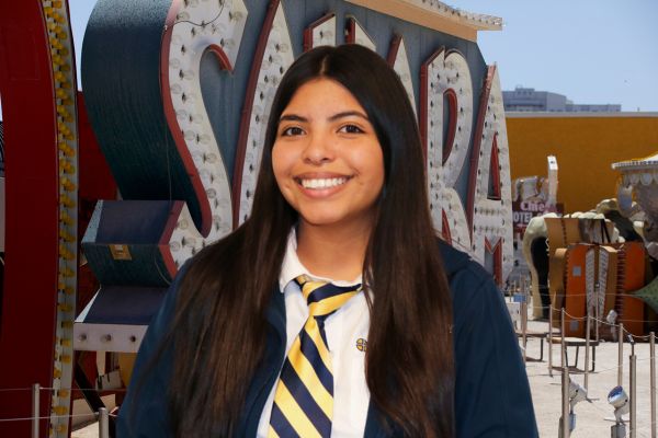 Mya Hernandez has long brown hair and is wearing a dark blue jacket, white button up shirt, and a blue and yellow tie. She is in front of the vintage Saraha sign which is dark teal, red, and white.