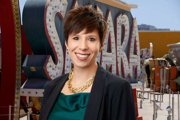Karen Alonso, with short brown hair, gold chain necklace, green silk top, and black blazer. The background image features the iconic Sahara Las Vegas Sign with dark teal, background, white letters, and a red outline.