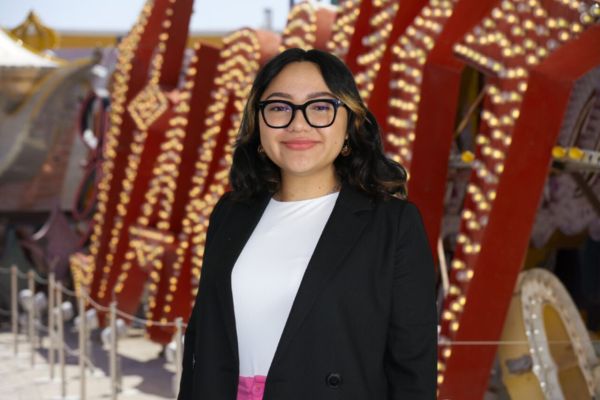 Karina Felix De La Riva has brown hair with black glasses. She is wearing a white blouse, black blazer, and pink trousers. The background behind Karina is the iconic Stardust sign from the Neon Museum and is red with gold lights.