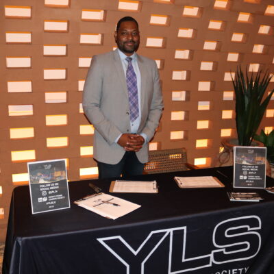Jason Williams in grey suit standing in front of Kassi Beach House entrances behind black table with YLS logo and flyers.