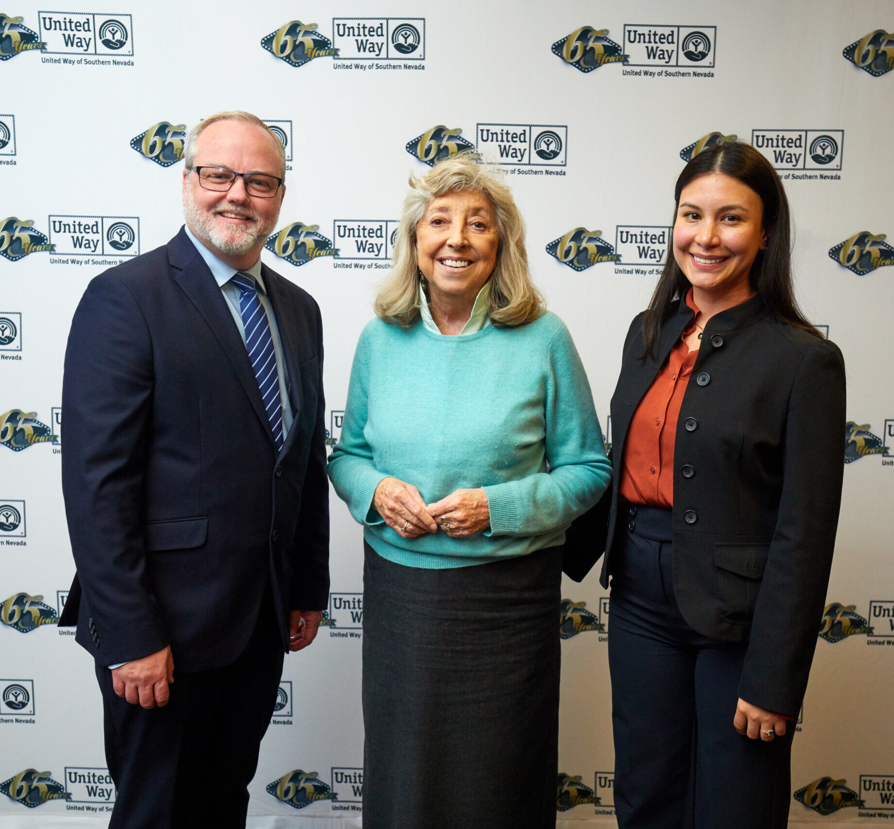 Samuel Rudd, Congresswoman Dina Titus and Janet Quintero standing in front of white background with black and gold UWSN 65th logo. Samuel and Janet are in black suits and Dina Titus is in a teal sweater.