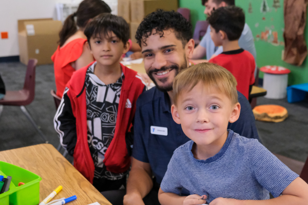 two children smiling at the camera at a worktable with a YMCA employee