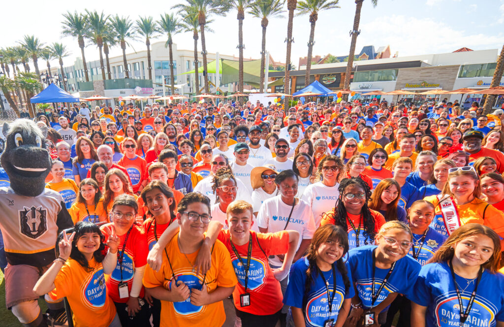A large, vibrant crowd of volunteers wearing colorful shirts gathers outdoors under palm trees during United Way of Southern Nevada’s Day of Caring event. Smiling participants in orange, blue, red, and white shirts celebrate together, with a mascot on the left side of the group.